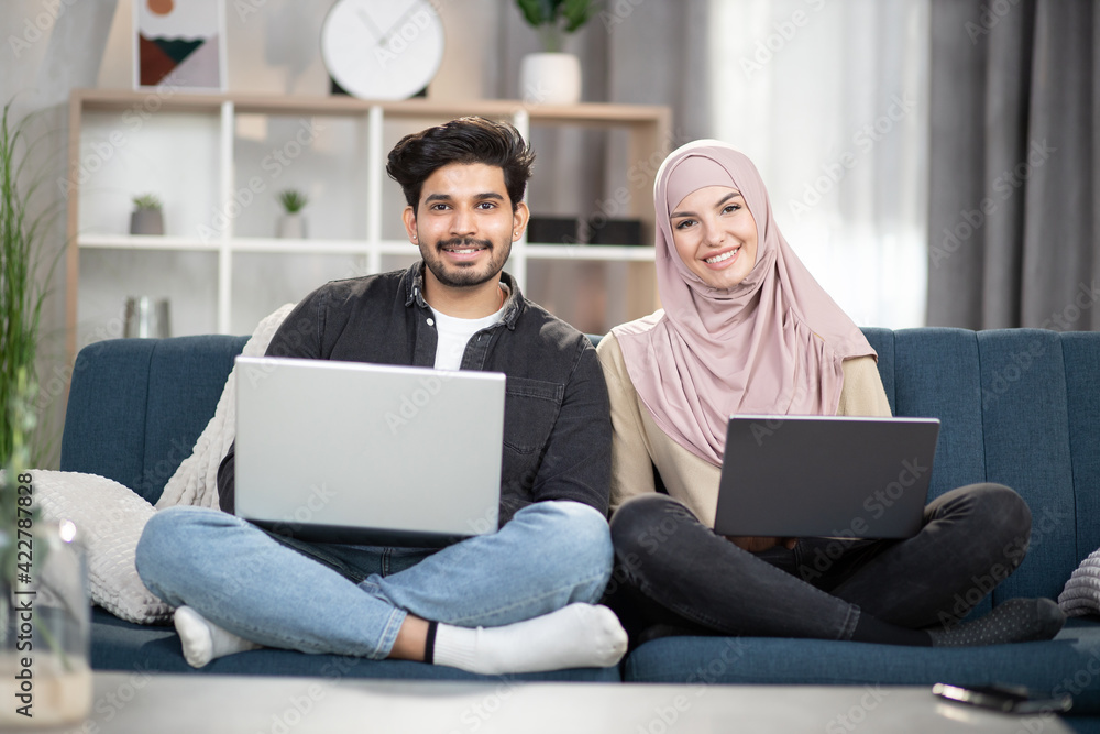 Young 30-aged smiling happy multiethnic couple, bearded Indian man and Muslim woman in hijab, sitting in lotus pose with laptops on comfortable couch at home while looking into camera.