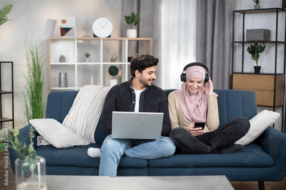 Distance learning and work. Leisure time with gadgets at home. Young 30-aged muslim male and female friends, sitting on sofa at cozy living room and using laptop, smartphone and earphones. Distance learning and work. Leisure time with gadgets at home. Young 30-aged muslim male and female friends, sitting on sofa at cozy living room and using laptop, smartphone and earphones.