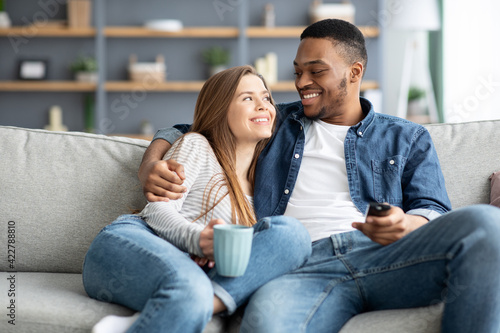 Happy loving interracial couple relaxing at home, watching tv and drinking coffee