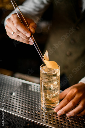 male bartender neatly decorates glass with cold drink by orange zest using tweezers