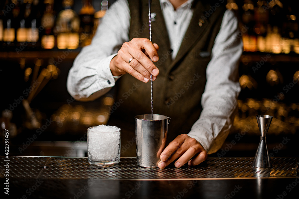 view on bartender holding long bar spoon and stirring cocktail in steel mixing cup