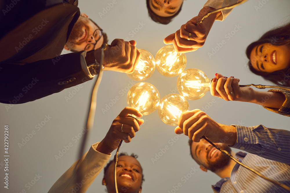 Bottom view of interracial colleagues stacking together light bulbs as a symbol of a new business idea. Concept of sharing ideas, business, teamwork and brainstorming. Selective focus. - obrazy, fototapety, plakaty 