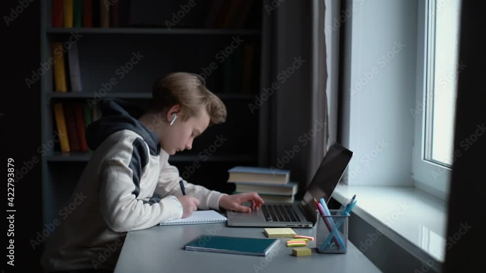 Focused pupil boy wearing wireless earphone learning online using laptop making notes in copybook sitting at desk at home, side view. Concept of distance education. Tracking shot in slow motion Focused pupil boy wearing wireless earphone learning online using laptop making notes in copybook sitting at desk at home, side view. Concept of distance education. Tracking shot in slow motion