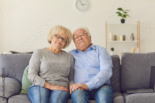 Smiling loving elderly couple wife and husband in jeans sitting on sofa, hugging and looking at camera together over room interior background. Elderly people active lifestyle and love concept