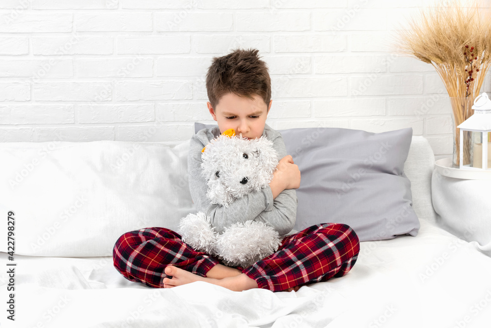 Obraz premium Angry boy in pajamas in bed. Child with a toy in bedroom at home in the morning. Sad schoolboy hugs teddy dog. Resentment, anger, annoyance, misunderstanding, bad mood, kids emotion, care, childhood