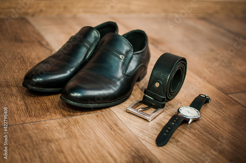 Black leather men's shoes, a watch and a belt lie on the floor. Groom accessories