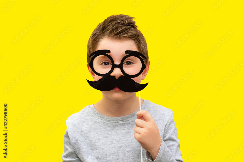 Funny happy boy holding black hipster mustache and glasses photo props, child pretends to be dad, professor or nerd on yellow background, Fathers Day concept, mens health, smart kids, joke, emotion Funny happy boy holding black hipster mustache and glasses photo props, child pretends to be dad, professor or nerd on yellow background, Fathers Day concept, mens health, smart kids, joke, emotion