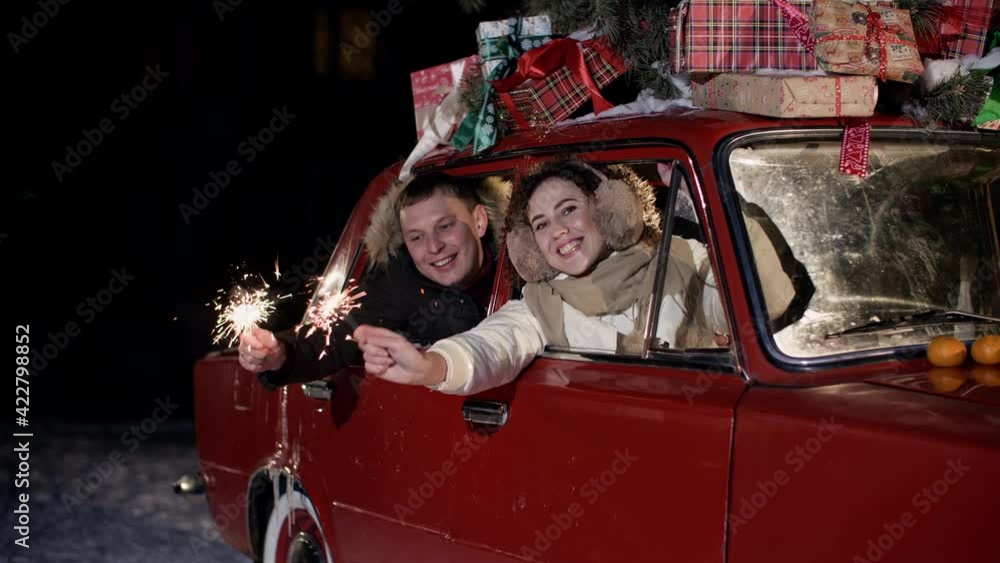 Happy couple holding sparklers and sitting in car with presents. Cheerful young couple holding burning sparklers and celebrating christmas in red vintage car with xmas gifts Happy couple holding sparklers and sitting in car with presents. Cheerful young couple holding burning sparklers and celebrating christmas in red vintage car with xmas gifts