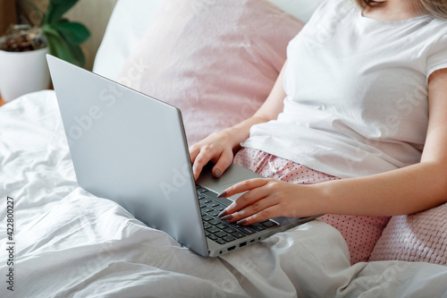 Female hands typing on laptop keyboard while lying in bed. Woman work using laptop while sitting in bed at morning home. Teen girl in pink pajamas studying online or planning her day.