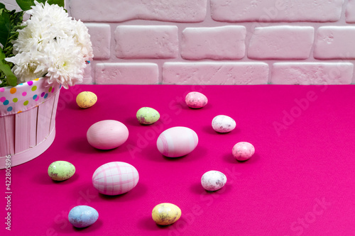Colorful Easter eggs and chrysanthemum flowers on a red table against a white brick wall background.