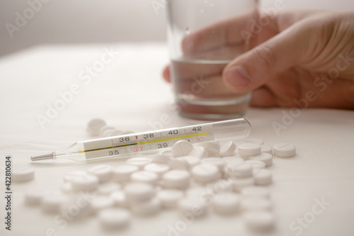Medical thermometer, with a temperature of 37 degrees, lies in a pile of pills, in the background a hand takes a glass of water. 