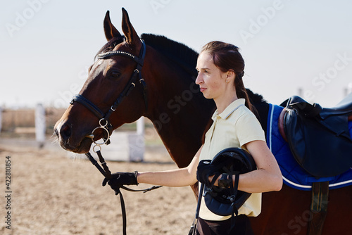 Young female farmer taking care about horse on ranch on summer day, copy space. Girl and brown horse in paddok outdoors photo