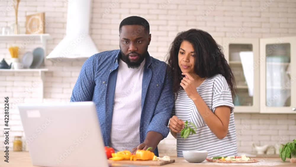 Portrait of young happy latin couple using laptop while cooking in kitchen at home, looking at the screen, watching culinary tutorial lesson, prepares their favorite recipe of healthy vegetable pizza Portrait of young happy latin couple using laptop while cooking in kitchen at home, looking at the screen, watching culinary tutorial lesson, prepares their favorite recipe of healthy vegetable pizza