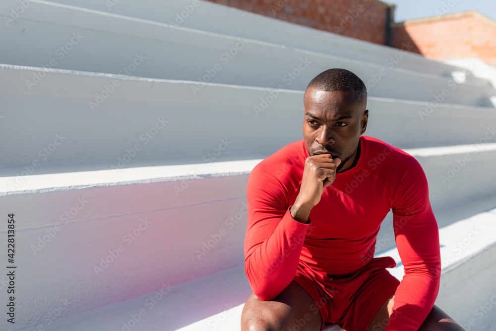 Fototapeta premium Athletic black man in sportswear sitting on stairways with joined hands looking away with serious determined face. copy space