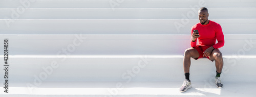 Horizontal banner black African man with sportswear sitting on white steps using smartphone. Minimalism and copy space.