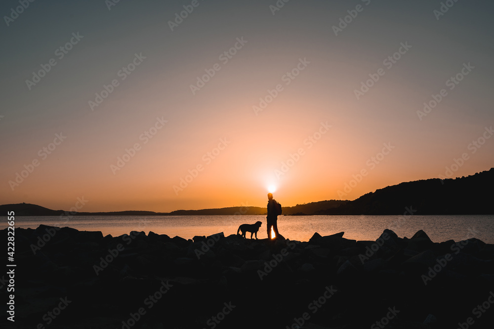 Silhouette of hiker man standing on the rocks in a coastline on the beach with his dog. Background with beautiful sunset. Silhouette of hiker man standing on the rocks in a coastline on the beach with his dog. Background with beautiful sunset.
