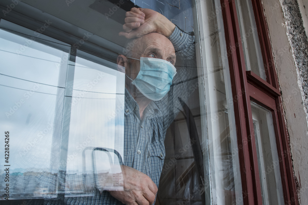 Senior sick man in medical face mask looking through big nursing house window.