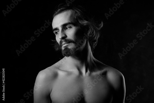Portrait of a handsome young man with a naked torso. A man with curly hair and a beard. Black and white photo. Muscle, fitness, dance. Body sculpture. Orientation.