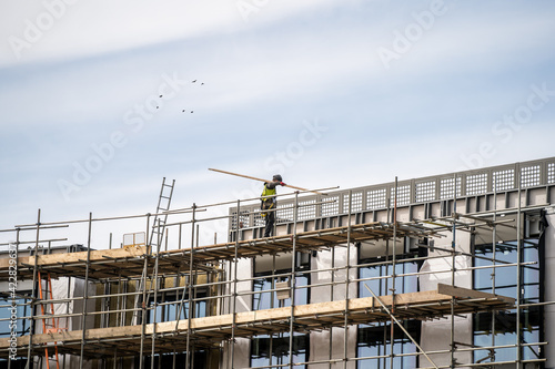 Anonymous scaffolder in PPE erecting framework of wooden planks and tall scaffolding poles high up on modern new building construction site. Professional dangerous job with safety equipment and tools.