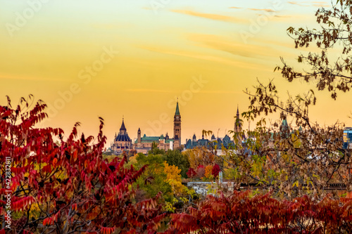 A brilliant yellow and orange sunset fills the sky around the Parliament Buildings, with red autumn foliage in the foreground