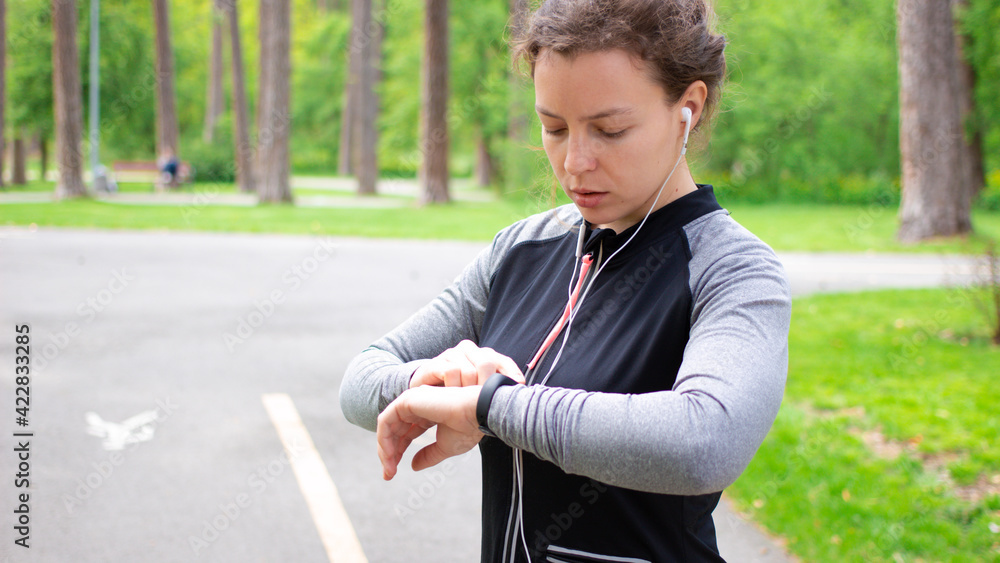 Athlete girl jogger adjusting fitness bracelet, using setting app smart watch to monitor her performance progress before workout, standing in green park outdoors. Checking pulse or sport information Athlete girl jogger adjusting fitness bracelet, using setting app smart watch to monitor her performance progress before workout, standing in green park outdoors. Checking pulse or sport information