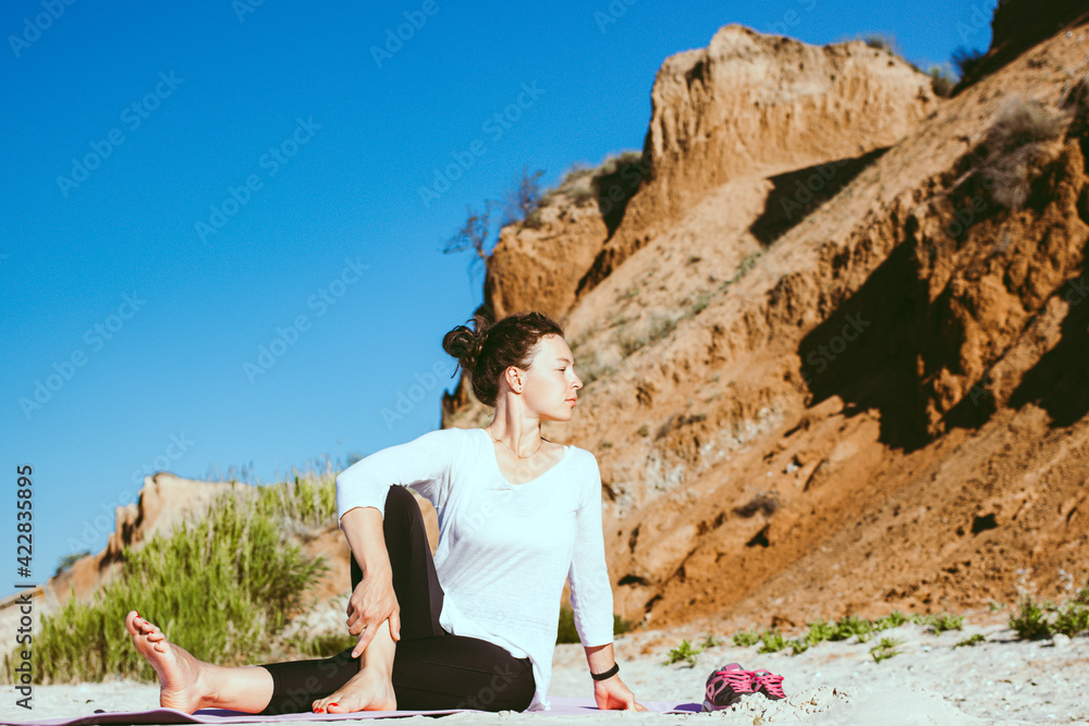 Seated spinal twist. Fit woman practicing yoga outdoor stretching lower back for spine health sitting on fitness mat on sandy beach with cliff landscape view. Training class on fresh air in summer