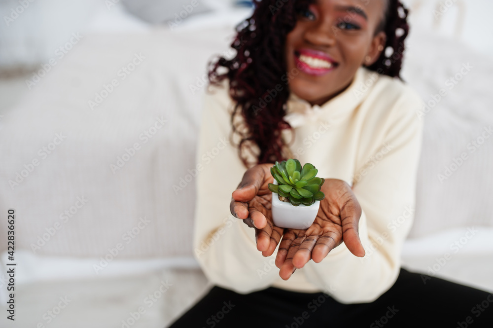 African woman in hoodie hold small flowerpot at home. African woman in hoodie hold small flowerpot at home.