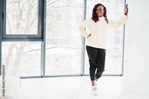 African woman in hoodie stand indoor apartment near large windows.