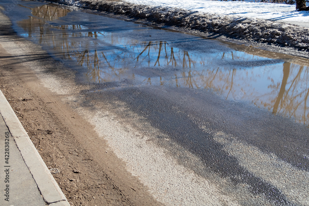 Dirty asphalt road with large puddles from melting snow and a layer of sand at the sidewalk. Consequences of de-icing urban roads in winter - dust and dirt from reagents and sand mixtures