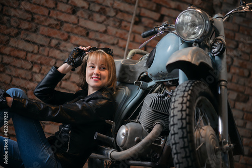 Young biker woman is sitting near the retro motorbike in the old garage and smiles.