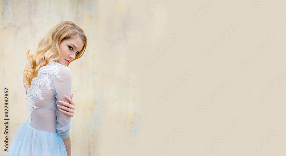Portrait of a young blonde woman in a delicate blue wedding dress against a beige wall. She turned her back