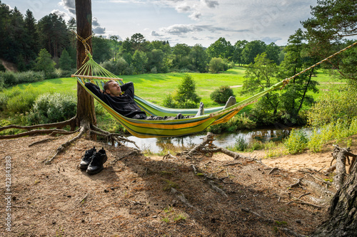 A man is resting in a hammock on the high bank of the river alone. His hiking boots stand side by side  photo