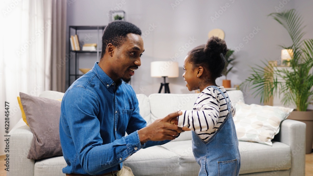 Side view of caring loving young happy dad speaking with cute daughter and hugging little girl at home, African American father embracing small child and smiling feeling love, portrait, family concept