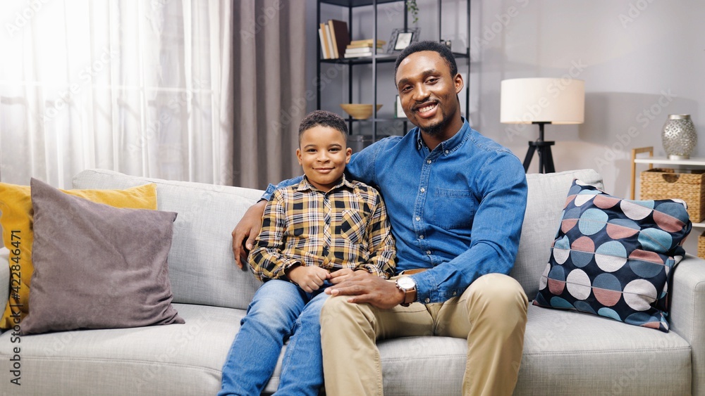 Portrait of happy young african american father and little son sitting at home on sofa and looking at camera smiling.