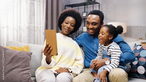 Happy young African American family sitting at home on couch with their little daughter and talking with relatives on video call waving their hands at tablet.