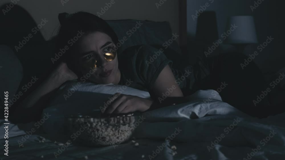 Night movie. Home leisure. Sleepless lifestyle. Relaxed curious woman with gold eye patches enjoying watching TV eating popcorn alone on bed late in dark bedroom with flicker blue light. Night movie. Home leisure. Sleepless lifestyle. Relaxed curious woman with gold eye patches enjoying watching TV eating popcorn alone on bed late in dark bedroom with flicker blue light.