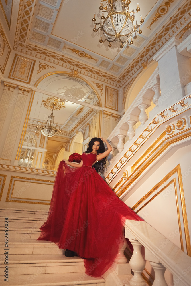 Beautiful brunette girl in a red ball gown on the background of white and gold palace interiors