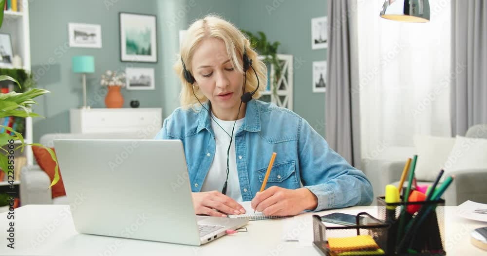 Portrait of Caucasian young pretty blonde woman in headset talking on webcam call on laptop studying online writing down information, female student having video lesson, webinar training concept Portrait of Caucasian young pretty blonde woman in headset talking on webcam call on laptop studying online writing down information, female student having video lesson, webinar training concept