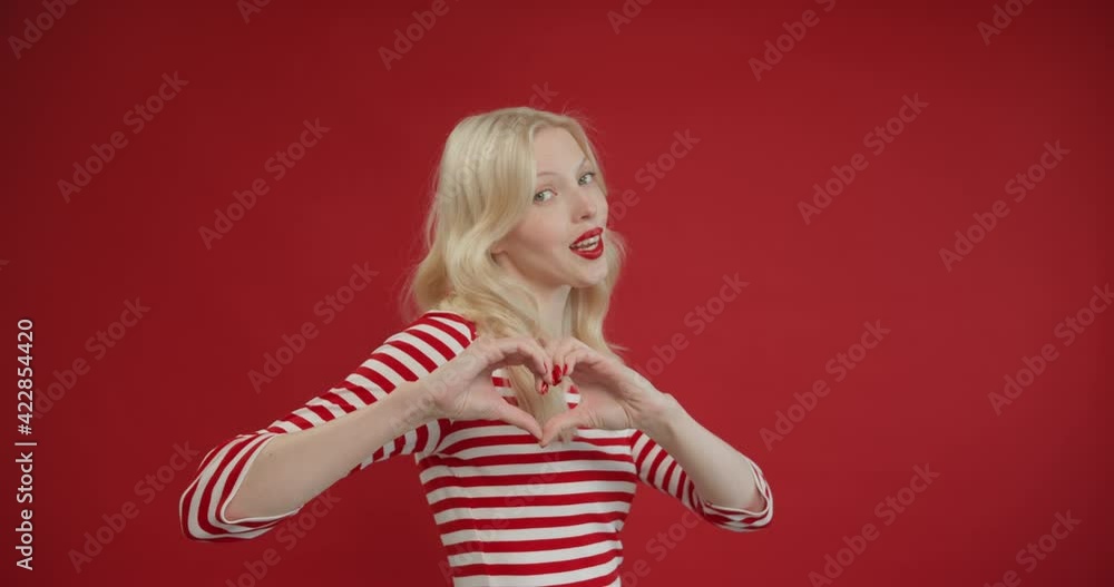 Close-up smiling young woman making sign heart shape by hand posing at red studio background. Close-up smiling young woman making sign heart shape by hand posing at red studio background.