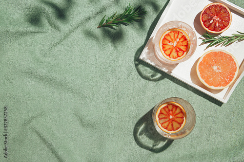 Summer scene with half of bloody orange and grapefruit,  glasses of water and rosemary with white wooden tray on pastel green beach towel. Drinks and refreshment concept. Sunlit flat lay. 