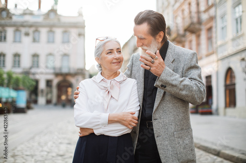 Smiling husband and wife in stylish clothes walking together on street. Happy senior couple hugging and chatting. Love in retirement.