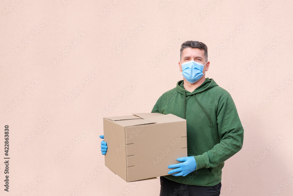 Middle-aged delivery man in medical mask and gloves holding and carrying a cardbox on pink wall background. Volunteer delivery of food or medicine during the coronavirus pandemic Middle-aged delivery man in medical mask and gloves holding and carrying a cardbox on pink wall background. Volunteer delivery of food or medicine during the coronavirus pandemic