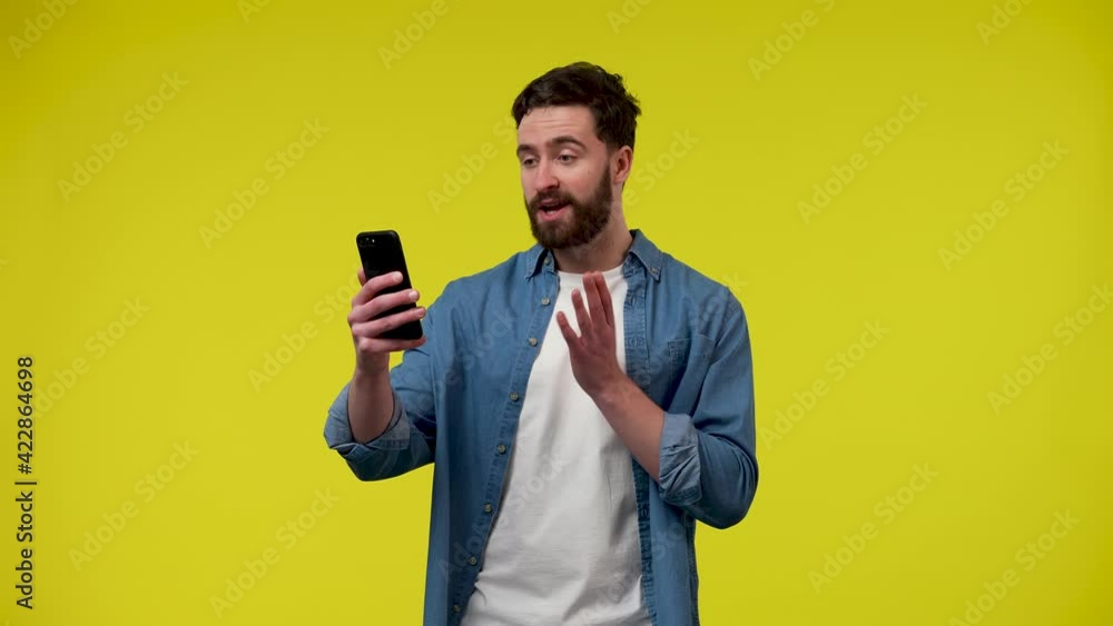 A man making a video call on his phone. The man waves his hand, greeting his interlocutor, points to the phone A man in the studio on a yellow background. Close up. Slow motion ready 59.97fps. A man making a video call on his phone. The man waves his hand, greeting his interlocutor, points to the phone A man in the studio on a yellow background. Close up. Slow motion ready 59.97fps.