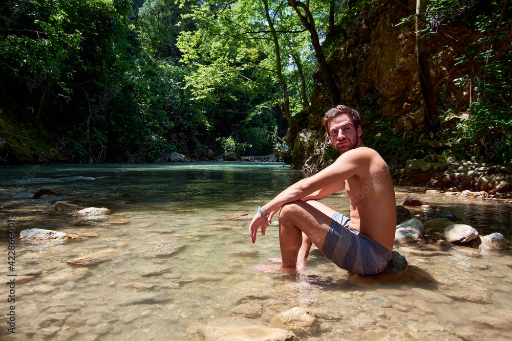 Fototapeta premium A handsome Caucasian man sitting on a rock in a river under sunlight