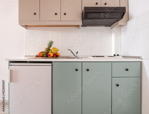 Closeup of white tile splashback on empty wall in simple cabinet module of modern interior kitchen with small fridge, vent, sink, stove, fruits plate