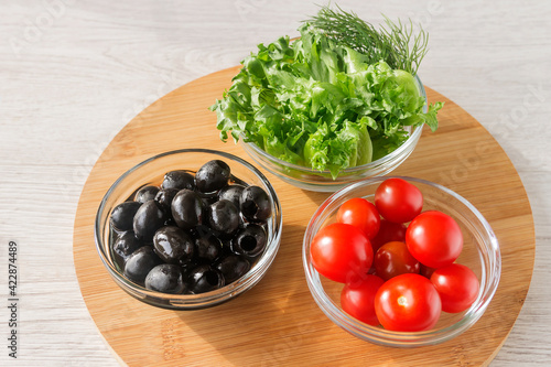 On a wooden kitchen board, three glass bowls with cherry tomatoes, olives, lettuce for a snack. 