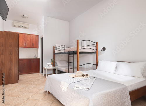 White modern interior of family suit bedroom with classic double bed, bunk bed with metal stairs, wooden kitchen cabinets, in single space of small room hotel studio apartment