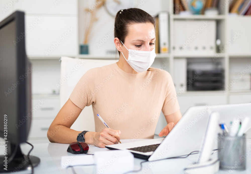 Woman in protective mask sitting at workplace with computer in her office