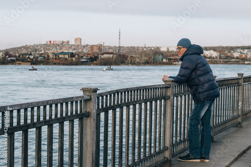 Canvas Print A middle-aged man in glasses, in a warm jacket and hat, stands on the embankment of the river against the backdrop of the cityscape