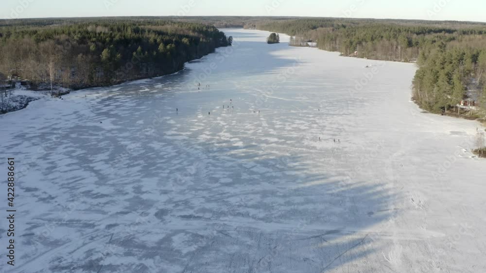 An aerial drone shot of a large frozen lake with a group of people ice skating on the surface - Daytime An aerial drone shot of a large frozen lake with a group of people ice skating on the surface - Daytime